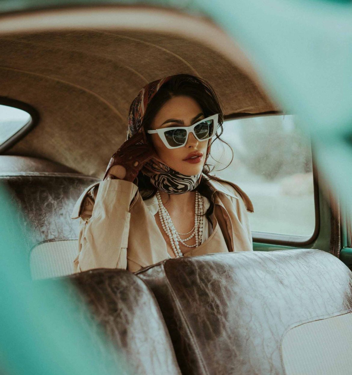 A woman wearing white cat-eye sunglasses, a beige trench coat, pearl necklaces and a printed scarf